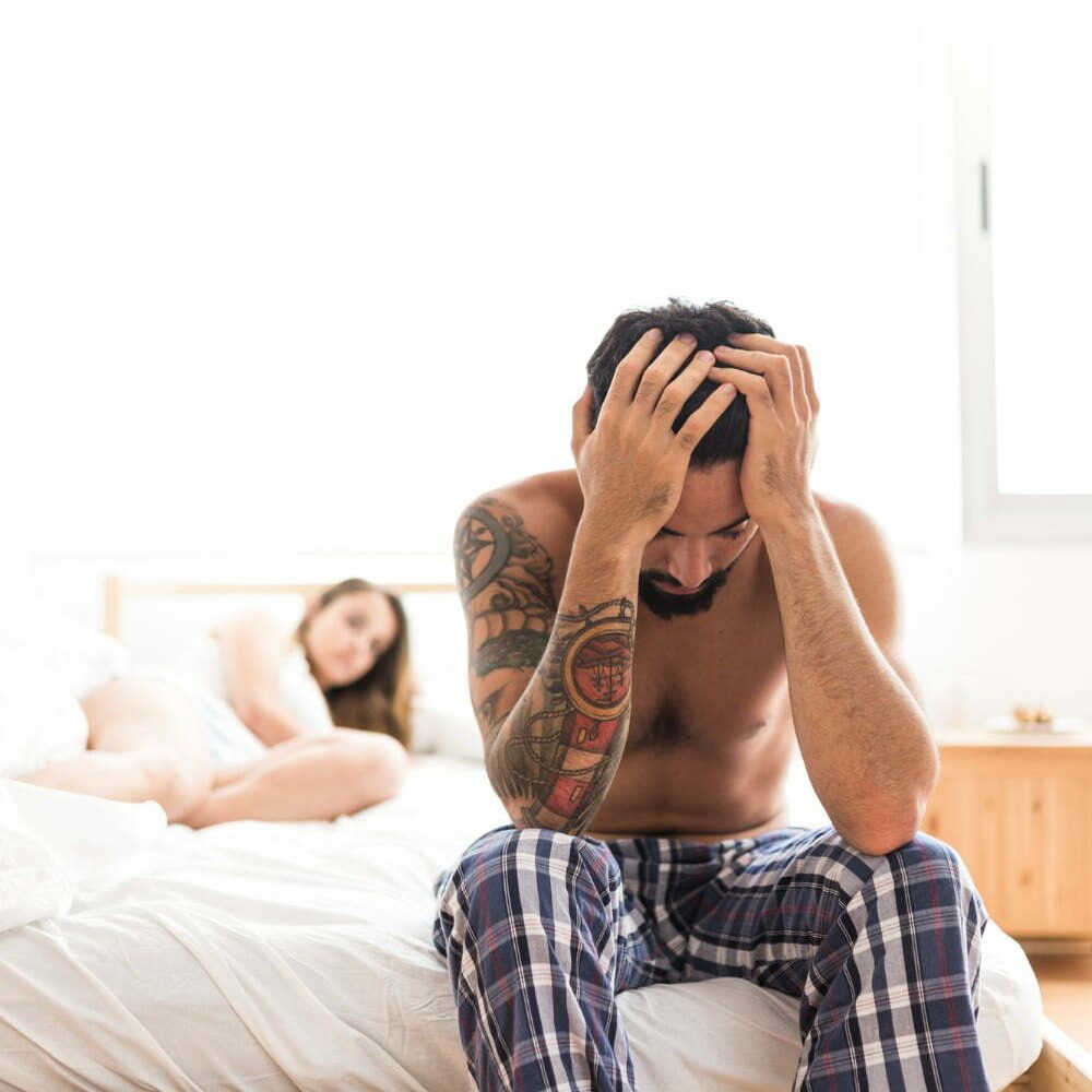 hombre sentado en la orilla de la cama, toc&aacute;ndose la cabeza, con su pareja detr&aacute;s, acostada en la cama. Problemas por trastono hipersexual