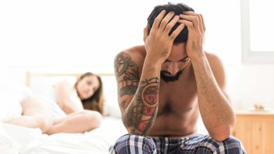 hombre sentado en la orilla de la cama, toc&aacute;ndose la cabeza, con su pareja detr&aacute;s, acostada en la cama. Problemas por trastono hipersexual