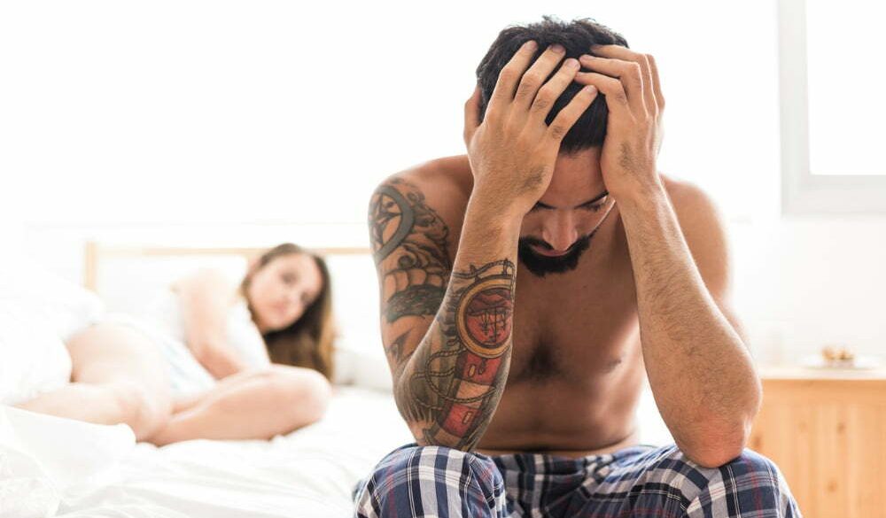 hombre sentado en la orilla de la cama, toc&aacute;ndose la cabeza, con su pareja detr&aacute;s, acostada en la cama. Problemas por trastono hipersexual