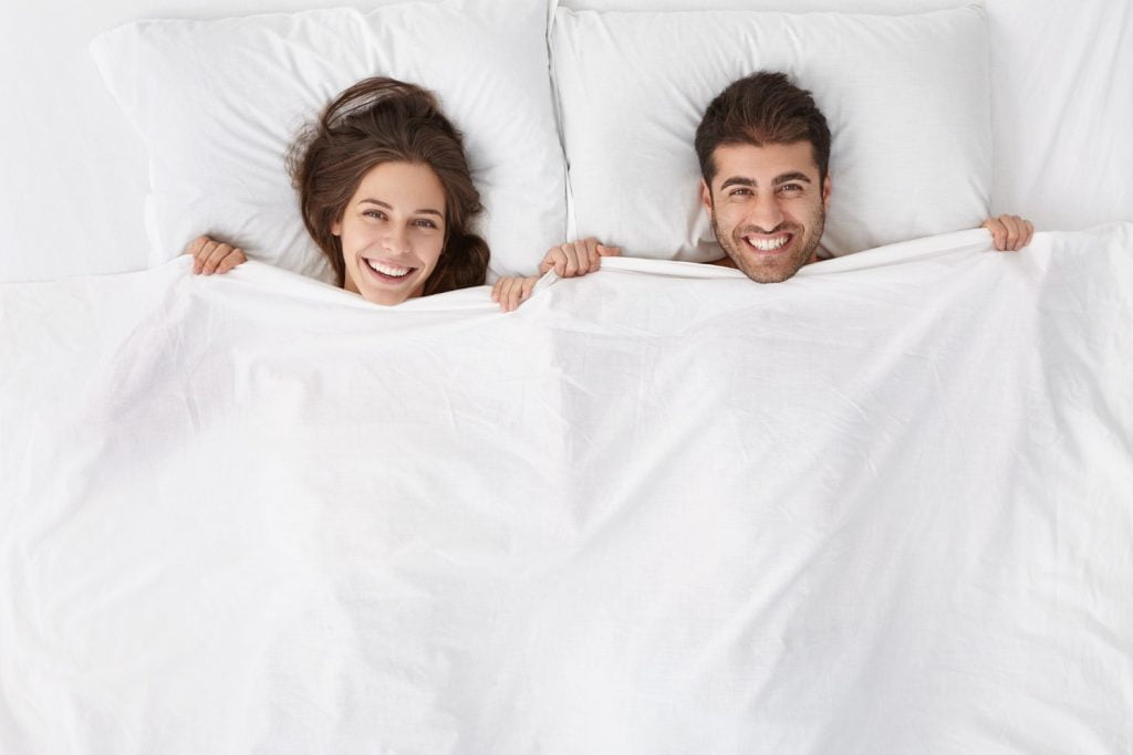 pareja acostada en la cama, tap&aacute;ndose con la s&aacute;bana hasta el cuelo, para dormir sin ropa