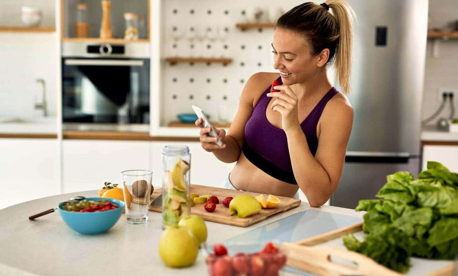 mujer con ropa de hacer ejercicio, comiendo frutas para perder peso, mientras mira su celular / Crisis de la mediana edad / masa muscular