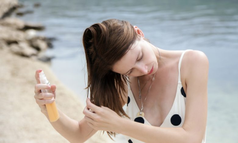 Mujer aplicando un spray de protección del cabello en la playa / cabello playa