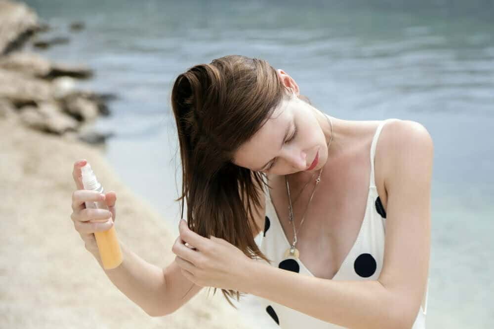 Mujer aplicando un spray de protecci&oacute;n del cabello en la playa / cabello playa
