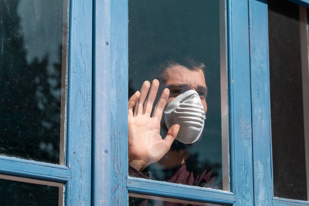 Hombre enfermo mirando por la ventana y usando máscara de protección / pandemia